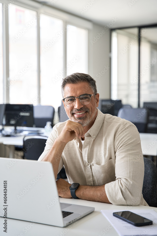 Vertical portrait of mature Indian or Latin business man ceo at table with laptop computer, working in modern office. Middle-age Hispanic smiling handsome businessman entrepreneur looking at camera. Vertical portrait of mature Indian or Latin business man ceo at table with laptop computer, working in modern office. Middle-age Hispanic smiling handsome businessman entrepreneur looking at camera.