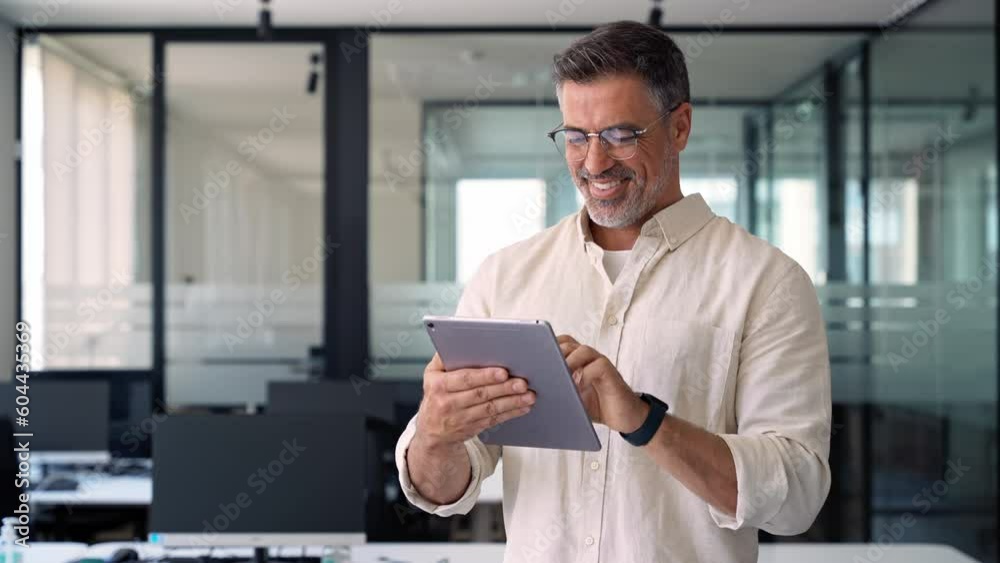 Happy Latin Hispanic bearded stylish mature adult professional business man, smiling Indian senior businessman CEO holding digital tablet using fintech tab application standing inside company office.