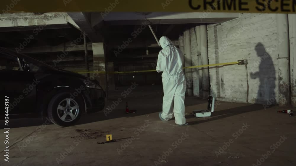 Full shot of unrecognizable male or female forensic investigator in white overalls walking around crime scene in dark parking, placing yellow markers and scale rulers next to blood stains and clues