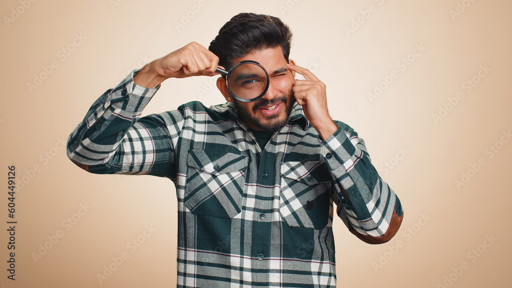 Investigator researcher scientist man holding magnifying glass near face, looking into camera with big zoomed funny eyes, searching, analysing. Handsome indian guy isolated on beige studio background Investigator researcher scientist man holding magnifying glass near face, looking into camera with big zoomed funny eyes, searching, analysing. Handsome indian guy isolated on beige studio background