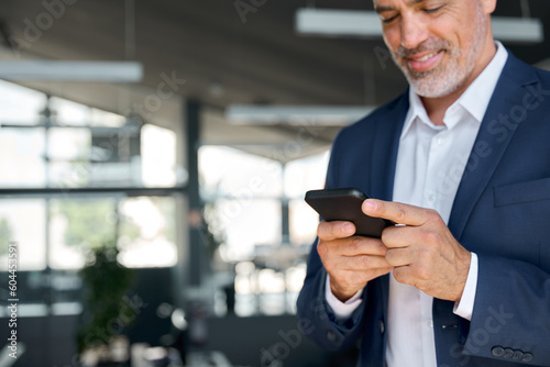 Smiling mid aged business man wearing suit standing in office using cell phone. Mature businessman professional executive holding smartphone in hand checking new mobile solution on cellphone. Close up