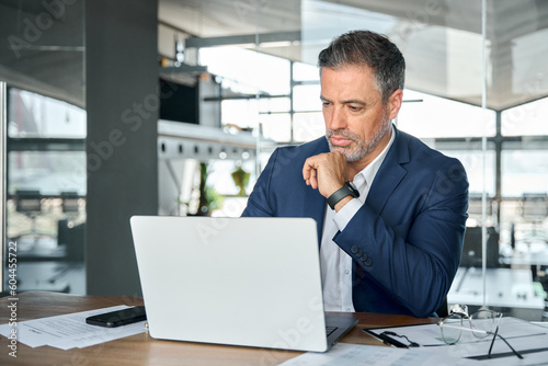 Busy serious mature business man ceo executive investor wearing suit looking at laptop computer analyzing financial data, managing corporate risks, thinking over project strategy working in office.