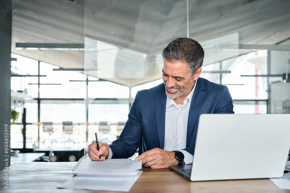 Happy mid aged professional business man executive ceo or lawyer wearing suit sitting at desk signing legal contract document putting written signature authorizing corporate deal in modern office. - obrazy, fototapety, plakaty 