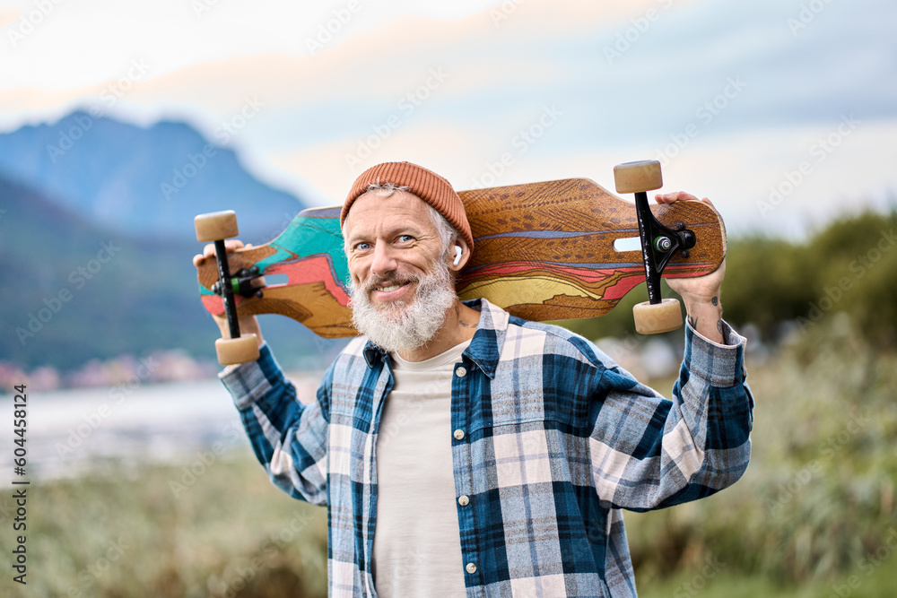 Active cool happy bearded old hipster man standing in nature park holding skateboard. Mature traveler skater enjoying freedom spirit and extreme sports hobby on mountains background. Portrait - obrazy, fototapety, plakaty 