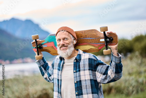 Active cool happy bearded old hipster man standing in nature park holding skateboard. Mature traveler skater enjoying freedom spirit and extreme sports hobby on mountains background. Portrait