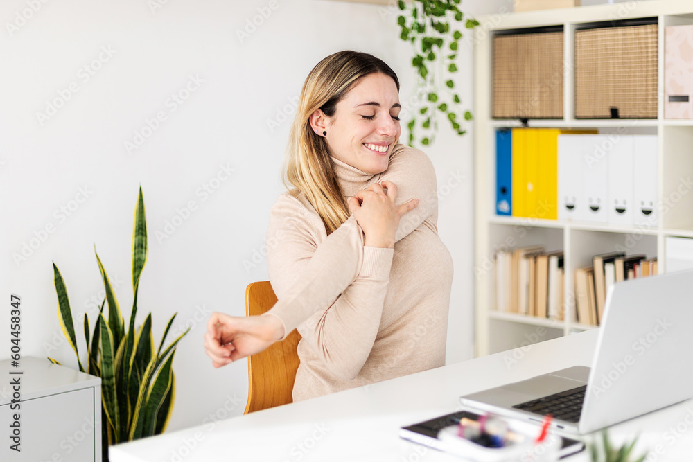 Young adult woman sitting on home office table stretching arms while working on laptop computer. Freelancer female worker taking break from work. Health concept - obrazy, fototapety, plakaty 