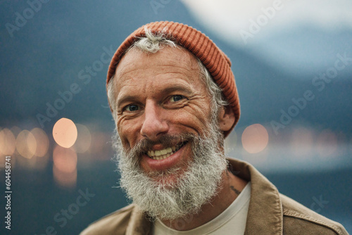 Happy older bearded man standing in nature park outdoors and laughing. Smiling active mature senior traveler looking at camera advertising camping tourism. Close up face front portrait.