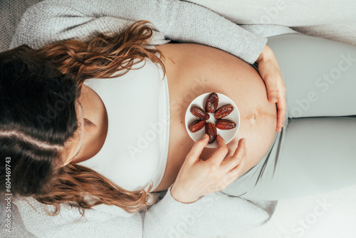 Top view of a woman sitting on the sofa eating Daddles while gently holding her belly in the last months of pregnancy. Pregnancy third trimester - week 34. top view. Sunny athmosphere.