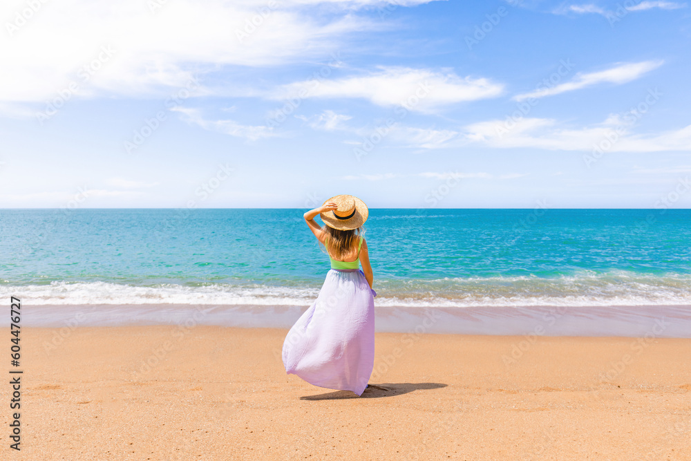 Cheerful woman in a straw hat standing alone on sandy beach, embracing the beauty and tranquility of tropical vacation. Clear ocean water with waves and sea breeze. Back view Cheerful woman in a straw hat standing alone on sandy beach, embracing the beauty and tranquility of tropical vacation. Clear ocean water with waves and sea breeze. Back view