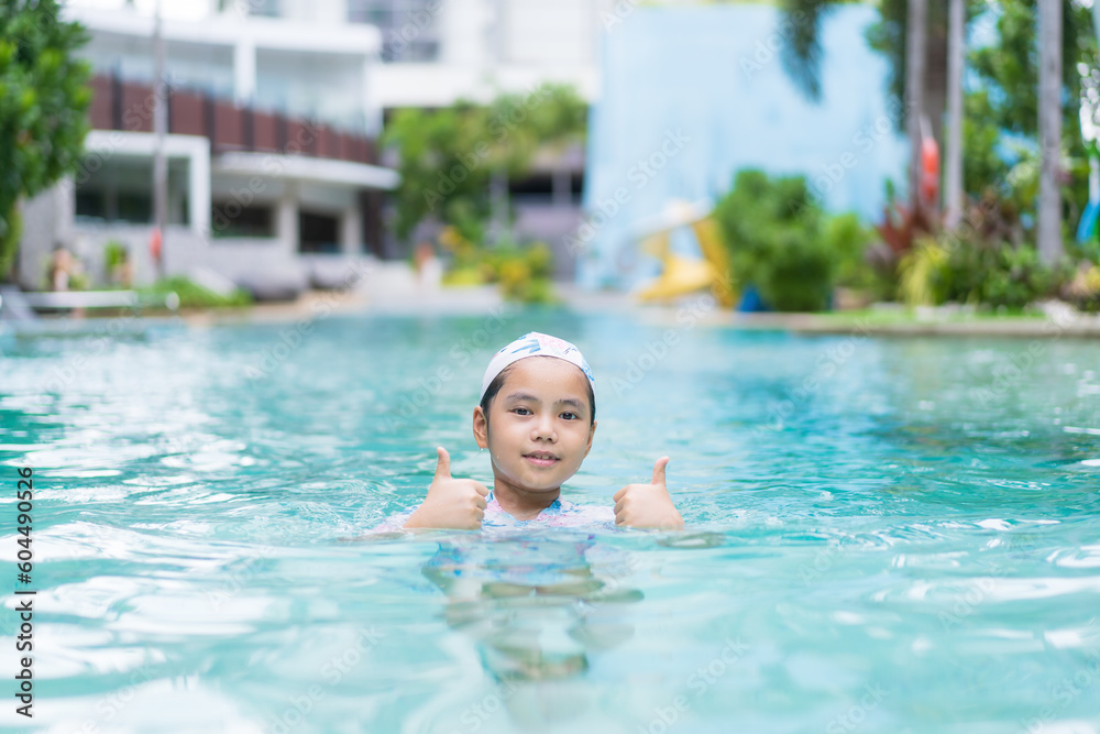 Asian child smile or kid girl wears cap swimsuit on swimming pool and happy fun to like thumbs up in water park or people learning swim to sports exercise on summer school or holiday vacation travel