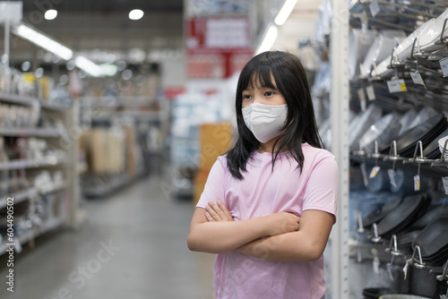 Asian child or kid girl wearing white face mask for close mouth nose to bored standing arms crossed flu cough sick to protect coronavirus covid-19 or pm 2.5 in shopping mall market or department store