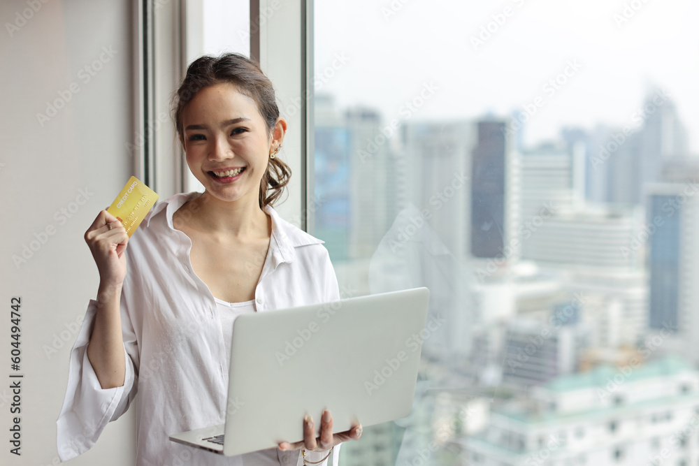 Fototapeta premium Asian woman in casual standing and holding credit card and using computer in house living room for online shopping payment. Confident businesswoman with city background. Lifestyle technology concept