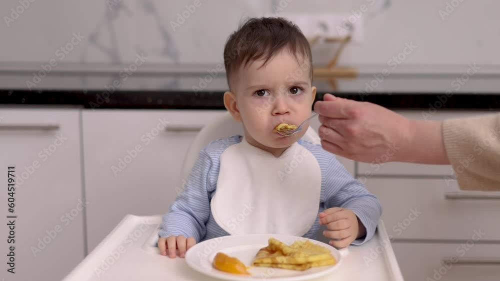 kid child eating pancakes with peach white bib writing in high chair,modern kitchen interior.mother mom woman hand feeding boy with fork.hungry upset toddler making air and graces,whims.crying face 4k kid child eating pancakes with peach white bib writing in high chair,modern kitchen interior.mother mom woman hand feeding boy with fork.hungry upset toddler making air and graces,whims.crying face 4k
