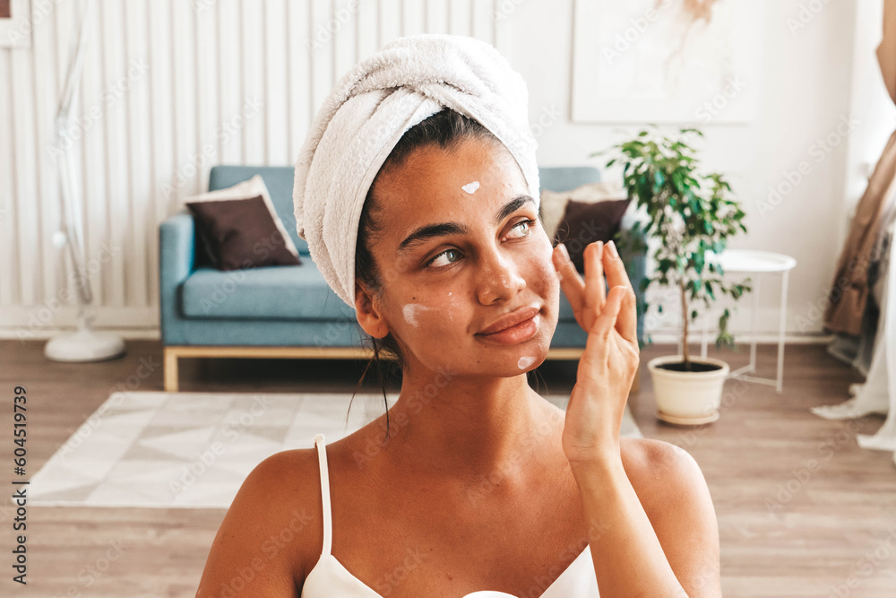 Young joyful charming brunette lady with towel wrapped on her head while sitting in living room, applying moisturizing face cream after bath procedures. Skin care and beauty concept Young joyful charming brunette lady with towel wrapped on her head while sitting in living room, applying moisturizing face cream after bath procedures. Skin care and beauty concept