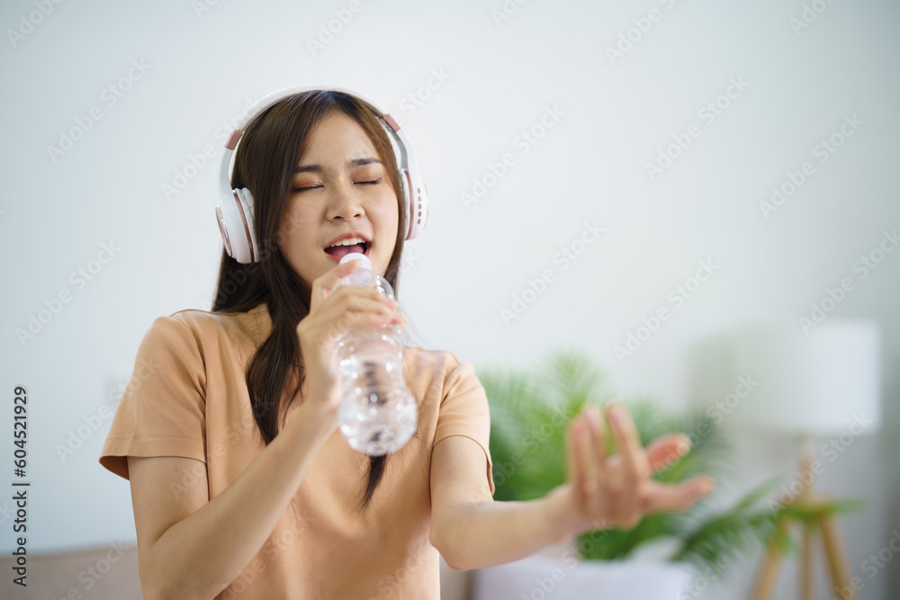 Happy Young Asian woman singing alone with bottle like microphone and wearing headphone love music happy relax in living room