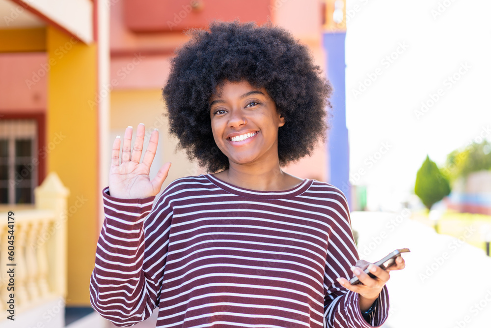 Fototapeta premium African American girl using mobile phone at outdoors saluting with hand with happy expression