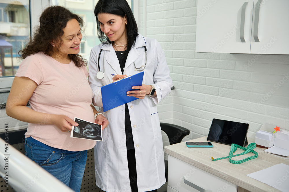 Obraz premium Multi ethnic happy positive pregnant woman holding the ultrasound scan of her baby, smiling while reading the prescription of her doctor gynecologist obstetrician during gynecological appointment