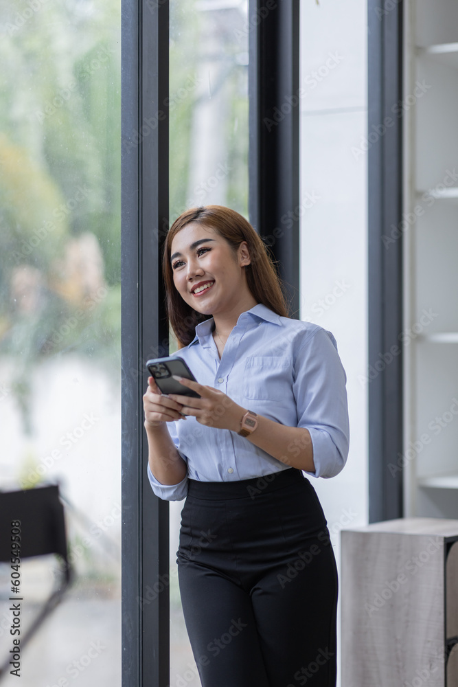 Smiling business Asian woman using her phone in the office. Small business entrepreneur looking at her mobile phone and smiling while communicating with her office colleagues Smiling business Asian woman using her phone in the office. Small business entrepreneur looking at her mobile phone and smiling while communicating with her office colleagues