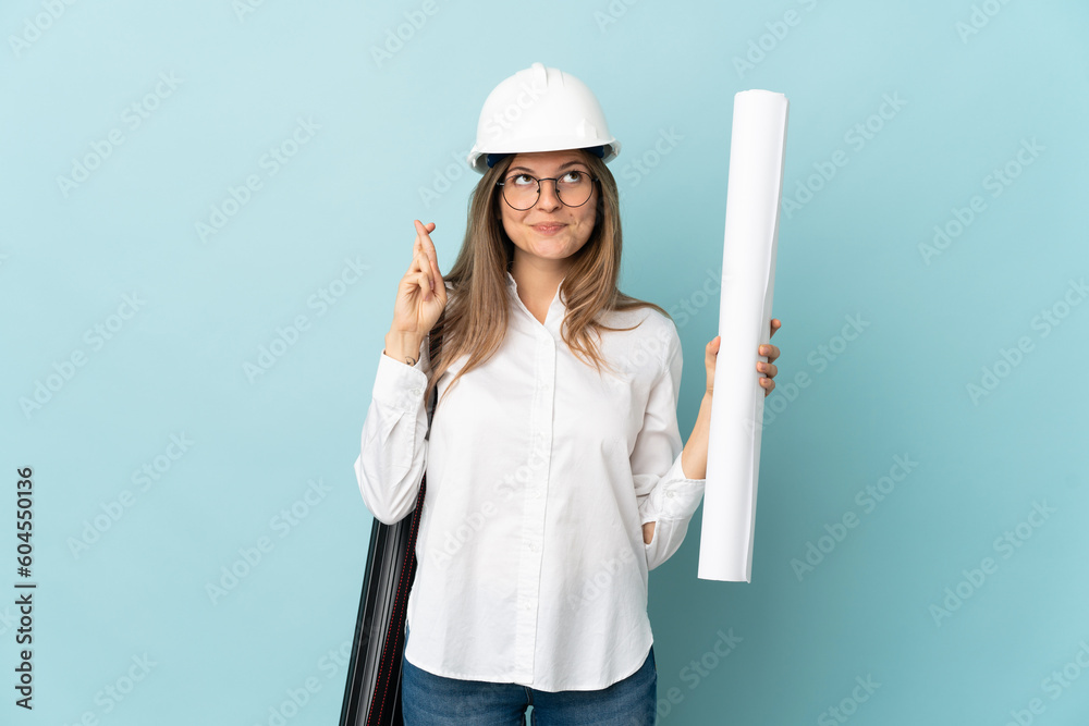 Slovak architect girl holding blueprints isolated on blue background with fingers crossing and wishing the best Slovak architect girl holding blueprints isolated on blue background with fingers crossing and wishing the best