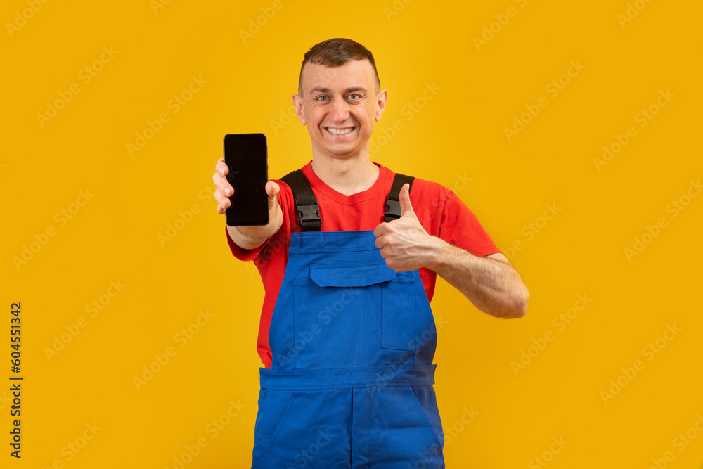 Young man in blue jumpsuit and red T-shirt holds smartphone with blank black screen and shows thumbs up. Copy space, mock up. Young man in blue jumpsuit and red T-shirt holds smartphone with blank black screen and shows thumbs up. Copy space, mock up.