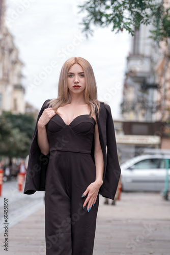 Elegant lady in business suit walks around the city. Young woman is blonde in black suit with deep neckline. Vertical frame.