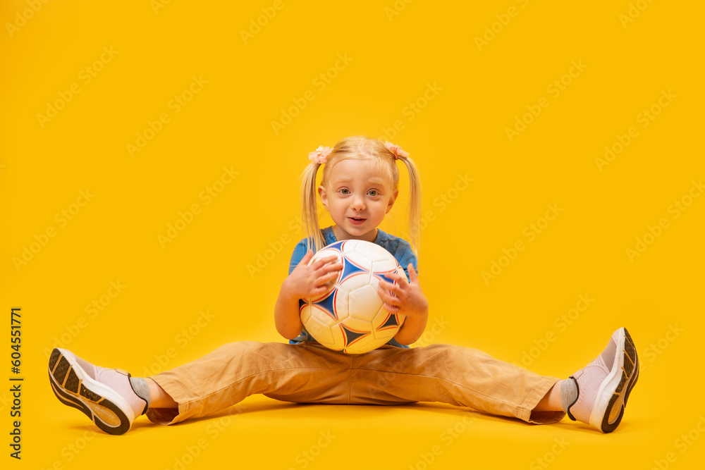 Little blonde girl with two tails sits on floor and holds soccer ball on yellow background. Womens childrens football. Little blonde girl with two tails sits on floor and holds soccer ball on yellow background. Womens childrens football.