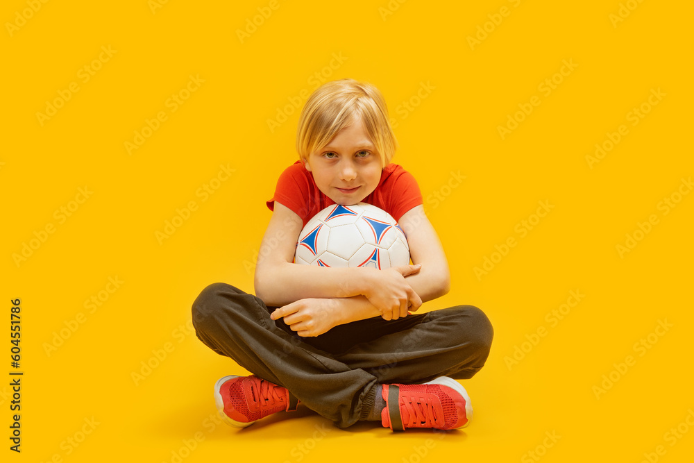 Portrait of boy sits cross-legged and hugs the ball in isolation on yellow background. Child footballer. Portrait of boy sits cross-legged and hugs the ball in isolation on yellow background. Child footballer.