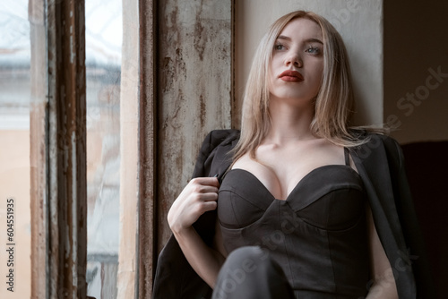 Young fair-haired woman wears black outfit bustier with sweetheart deep neckline sitting on the windowsill. Sad girl