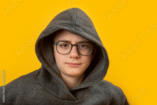 Portrait of boy with glasses wears hoodie and hood on yellow background. Shy unsociable teenager.