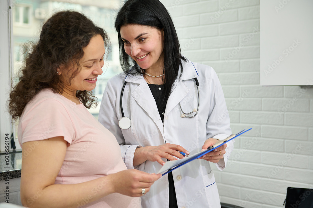 Charming confident female doctor wearing white medical gown, holding clipboard with medical documentation and consulting her new pregnant patient in a gynecological clinic. Obstetrics and gynecology Charming confident female doctor wearing white medical gown, holding clipboard with medical documentation and consulting her new pregnant patient in a gynecological clinic. Obstetrics and gynecology