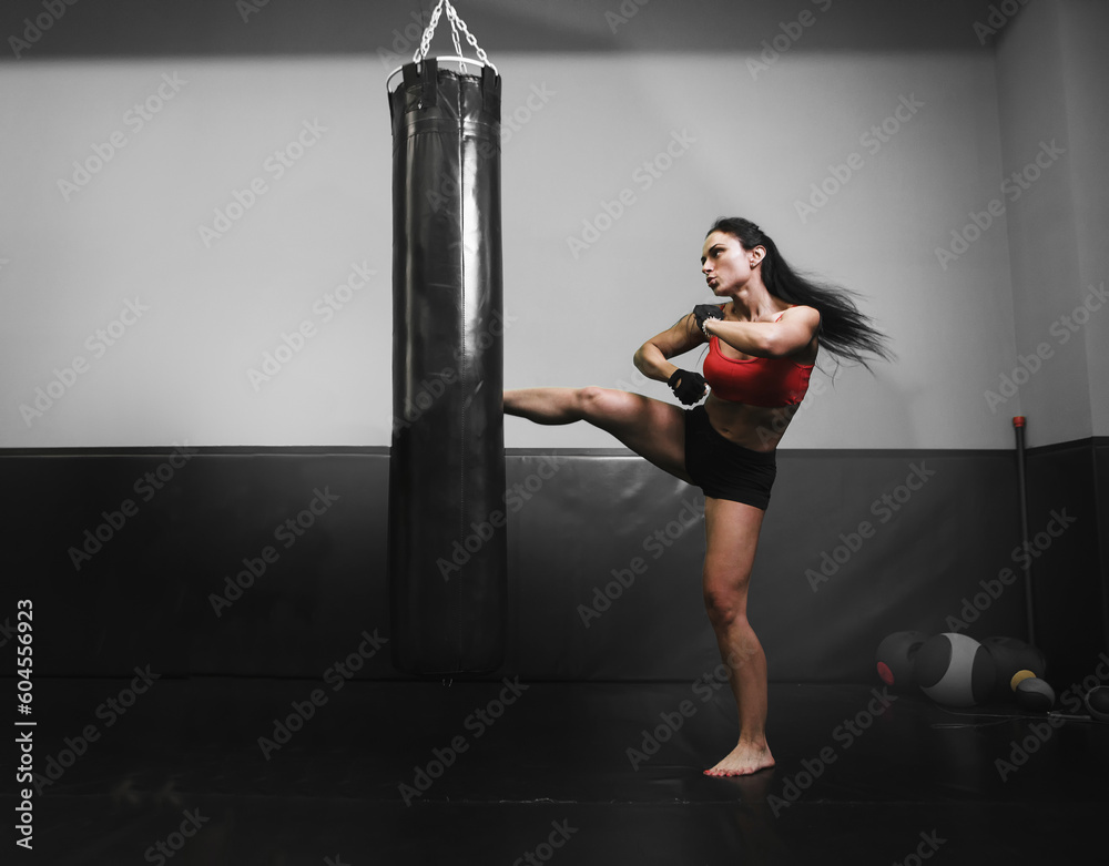 Caucasian woman in sportswear and with mma gloves kicking bag in the gym. Full length.
