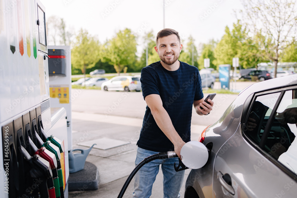 Bearded man refuelling car on gas station and looking into his smartphone. Man compares fuel prices Bearded man refuelling car on gas station and looking into his smartphone. Man compares fuel prices