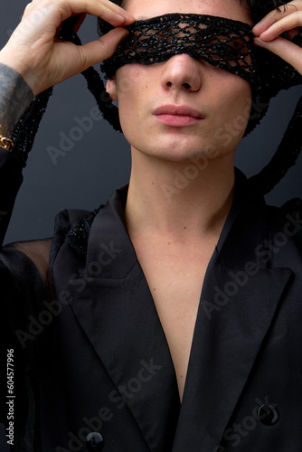 Close-up portrait of young sexy womanlike male in black suit with eye mask, isolated on black studio background. Passionate man is looking fashionable, stylish. fashion, beauty, people concept