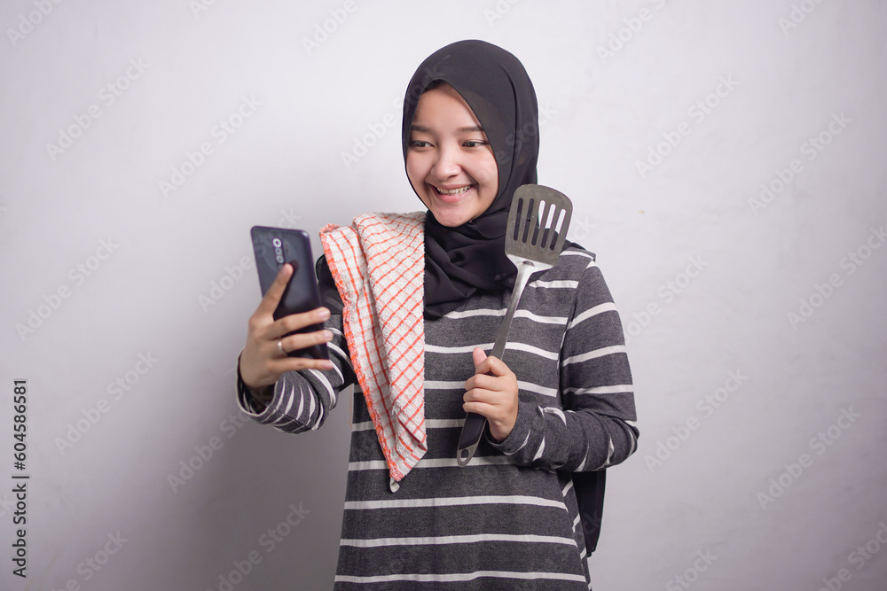 Portrait of smiling young asian woman wearing microfiber rag in black and white sweater and hijab holding mobile phone and spatula, showing blank phone isolated over white background.