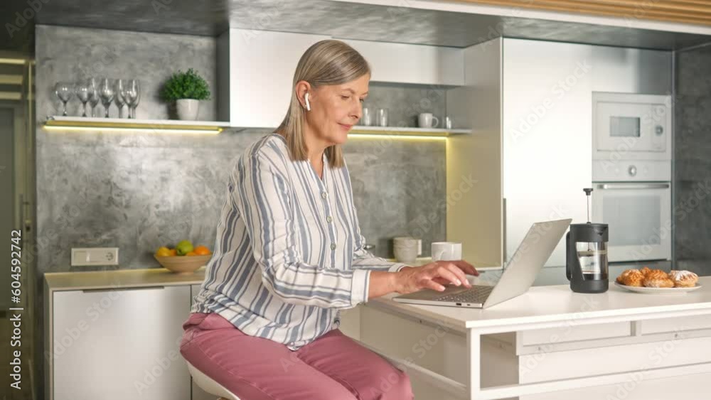 Charming Caucasian woman using technology devices while sitting on chair in kitchen. Busy female with earpods typing text on laptop and listening music. Remotely communicating at house. Charming Caucasian woman using technology devices while sitting on chair in kitchen. Busy female with earpods typing text on laptop and listening music. Remotely communicating at house.