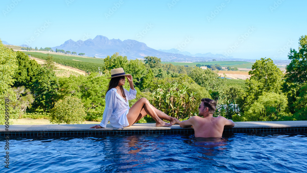Fototapeta premium Couple of men and women relaxing at a swimming pool with a view over a Vineyard landscape at sunset with mountains in Stellenbosch, near Cape Town, South Africa. 