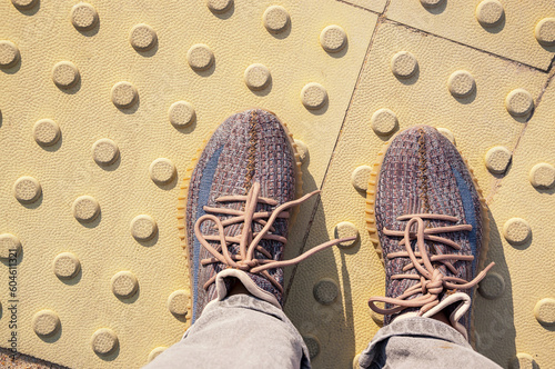 Women's feet in sneakers stand on a tactile tile. The road for the blind.