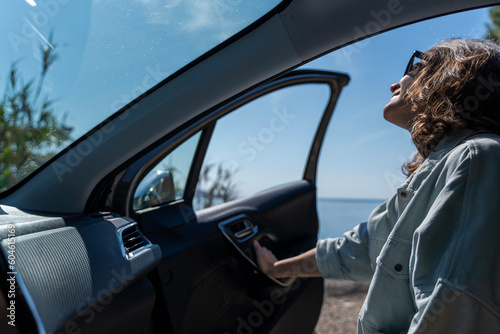 Young happy woman traveler opening car door at seaside. Summer vacation and auto travel concept