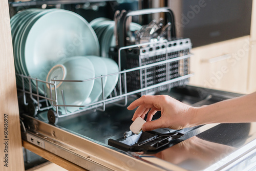 Female hand inserting dishwasher tablet into open built-in dishwasher machine photo