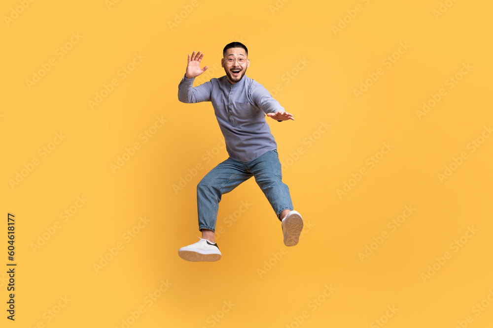 Carefree asian man in casual outfit jumping in air over yellow background