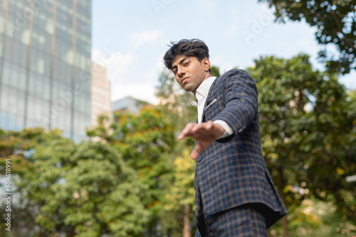 A stylish guy in a blue checkered suit in a serious face with a reaching his hand down gesture while standing outside, in the city. Sky, building and trees are shown in the background.
