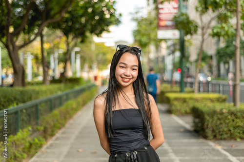 A pretty young lady standing in a park wearing a black strapless tank top and sunglasses, smiling prettily while her hands are behind her back. Trees,bushes and fence are in the background. © Mdv Edwards