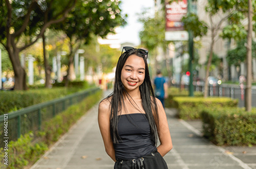 A pretty young lady standing in a park wearing a black strapless tank top and sunglasses, smiling prettily while her hands are behind her back. Trees,bushes and fence are in the background. photo