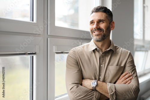 Positive middle aged businessman standing near window in company office, looking away and smiling, copy space