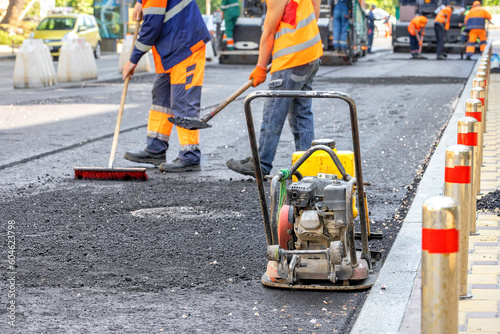 An old worn-out small compacting slab against the backdrop of a team of road workers laying fresh asphalt on a city street in blur.