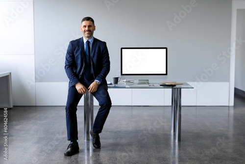 Middle aged businessman in formal suit sitting near office table with blank computer monitor, mockup for your design