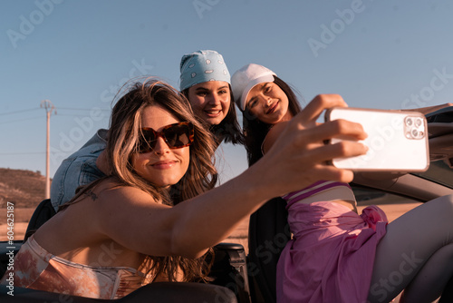 During the summer trip, the three young friends take a selfie while smiling. They are traveling in a convertible car.