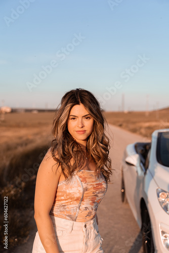 The white-skinned, wavy-haired young woman is in the countryside on a summer afternoon with her car