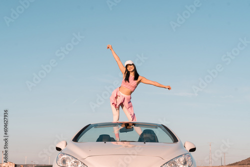 the white European girl dressed in summer clothes is perched on the seat of her white convertible car and raises her hands to the sky as she rides on a summer afternoon.