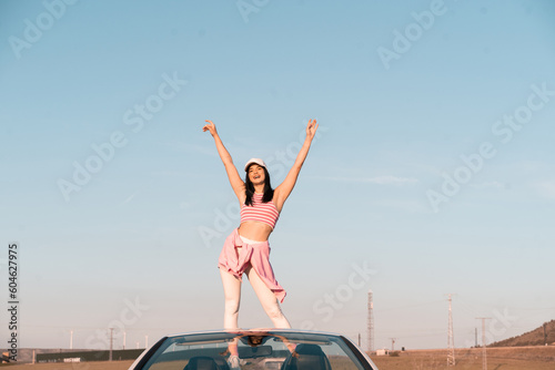 white European girl dressed in summer clothes sits on the seat of her white convertible car and raises her hands to the sky as she rides on a summer afternoon. © Alberto Cotilla
